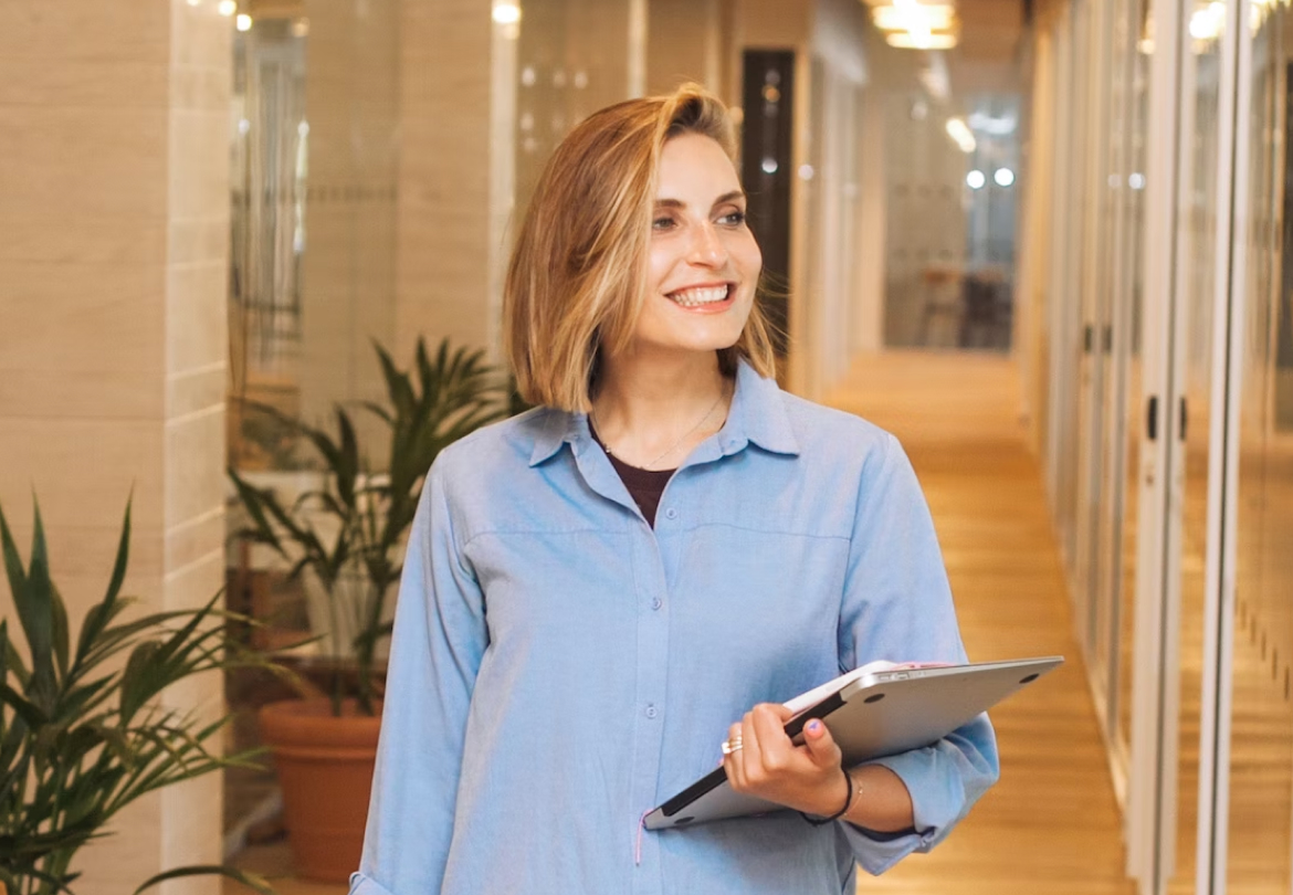 Woman in blue shirt holding a tablet, smiling indoors.