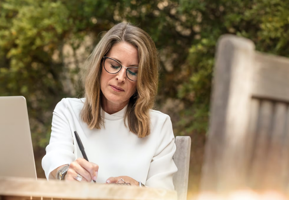 A woman in glasses writing at a table outdoors.