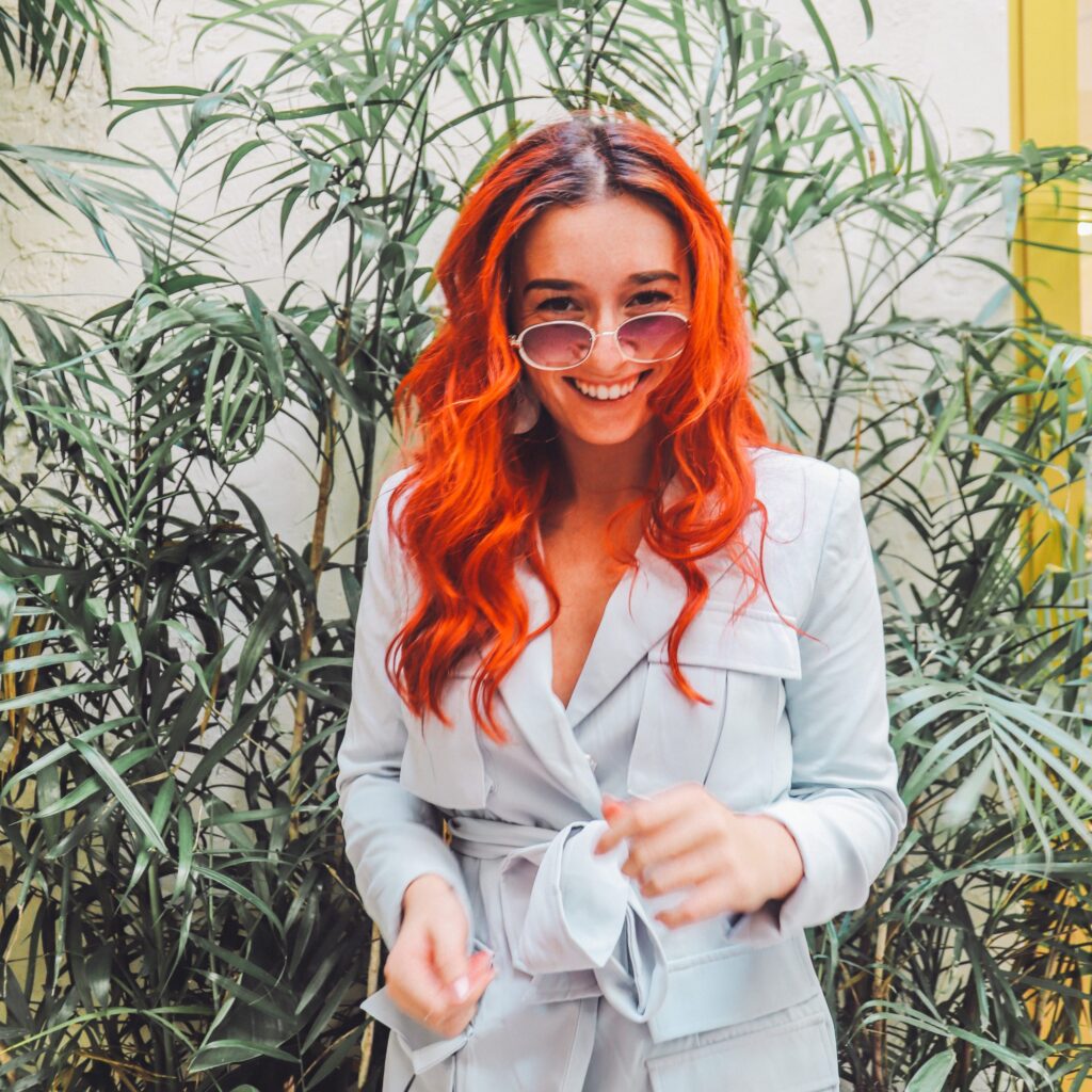 Woman with vibrant red hair and glasses stands amid greenery.